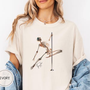 May include: Ivory-colored t-shirt featuring a skeleton figure pole dancing. The skeleton is brown and tan, with the name "Taylor" in cursive script. The shirt has a relaxed fit and rolled sleeves. The word "IVORY" is printed on a white circle.