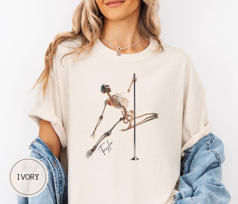 May include: Ivory-colored t-shirt featuring a skeleton figure pole dancing. The skeleton is brown and tan, with the name "Taylor" in cursive script. The shirt has a relaxed fit and rolled sleeves. The word "IVORY" is printed on a white circle.