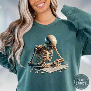 May include: Teal-colored sweatshirt featuring a detailed illustration of a skeleton assembling a jigsaw puzzle. The skeleton's head is a puzzle piece. The sweatshirt has a round neck and long sleeves. The text "BLUE SPRUCE" is in a circle on the bottom right.