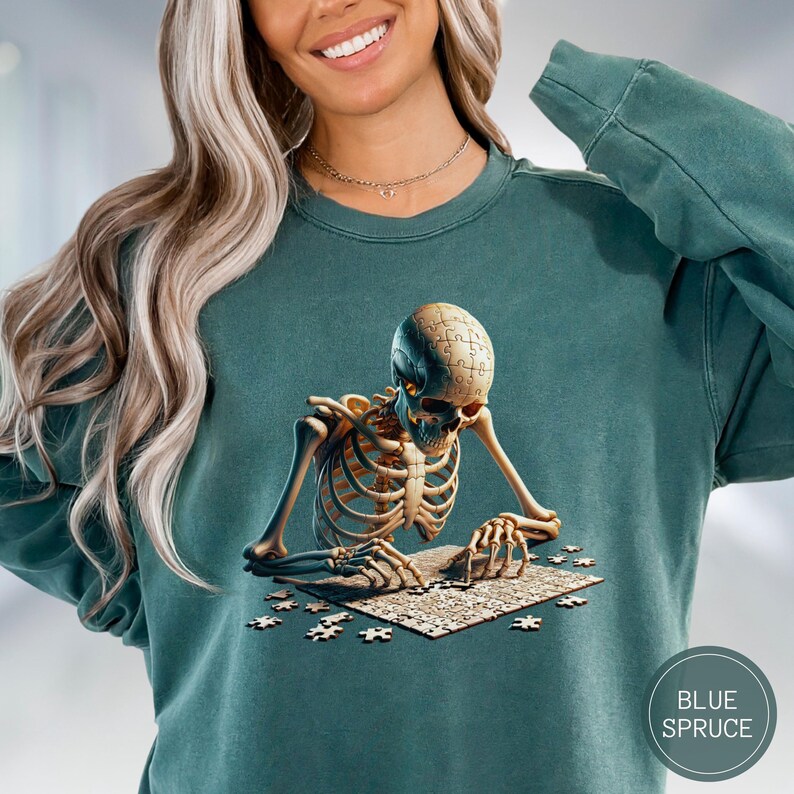 May include: Teal-colored sweatshirt featuring a detailed illustration of a skeleton assembling a jigsaw puzzle. The skeleton's head is a puzzle piece. The sweatshirt has a round neck and long sleeves. The text "BLUE SPRUCE" is in a circle on the bottom right.