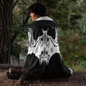 May include: A black hooded cape with a white bat skeleton design. The cape is worn by a person standing in a forest setting.