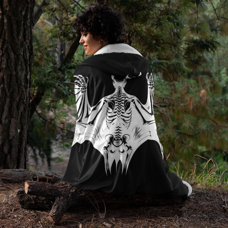 May include: A black hooded cape with a white bat skeleton design. The cape is worn by a person standing in a forest setting.