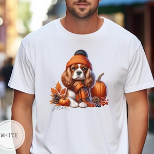 May include: White t-shirt with a graphic of a Cavalier King Charles Spaniel wearing a brown and orange hat and scarf, surrounded by fall leaves, pumpkins, and berries. The text "sam" is printed below the dog.