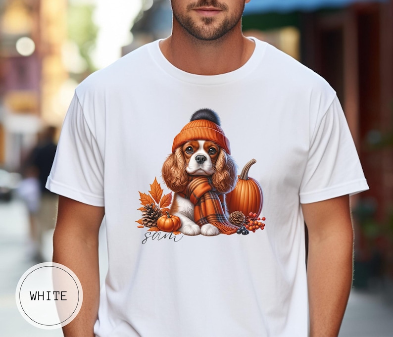 May include: White t-shirt with a graphic of a Cavalier King Charles Spaniel wearing a brown and orange hat and scarf, surrounded by fall leaves, pumpkins, and berries. The text "sam" is printed below the dog.
