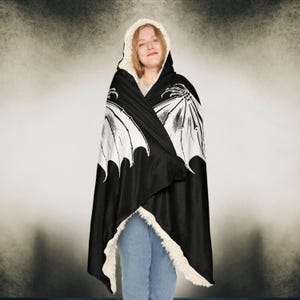 May include: A black hooded blanket with a white skeletal wing design. The blanket is made of soft fleece and has a sherpa lining.