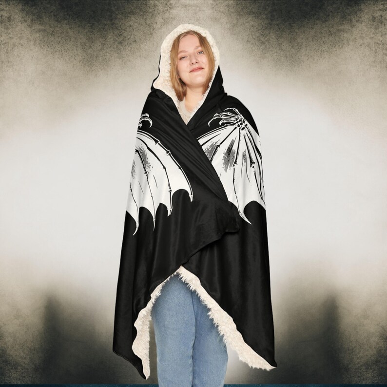 May include: A black hooded blanket with a white skeletal wing design. The blanket is made of soft fleece and has a sherpa lining.