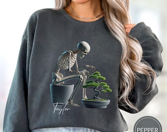 May include: Dark gray sweatshirt featuring a skeleton tending a bonsai tree in a pot. The skeleton is seated, focused on the bonsai. The name "Taylor" is written in cursive. A circular "Pepper" logo is in the bottom right.