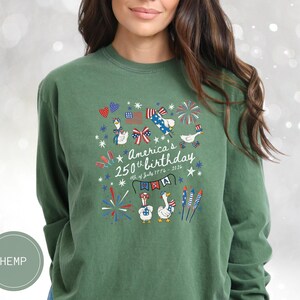 May include: Green long-sleeved top featuring a graphic commemorating America's 250th birthday. The design includes geese, fireworks, the American flag, and the text "America's 250th birthday 4th of July 1776-2026".
