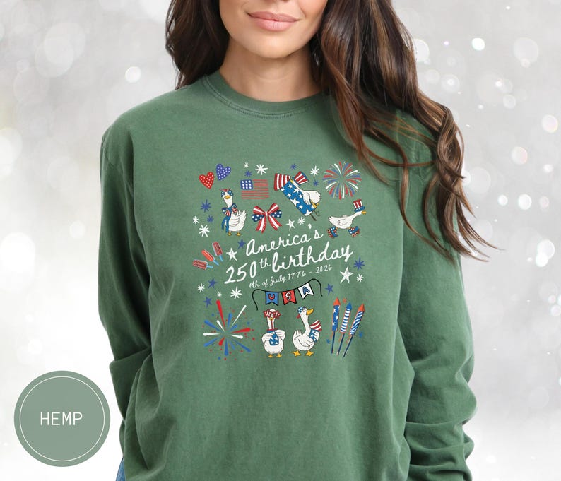 May include: Green long-sleeved top featuring a graphic commemorating America's 250th birthday. The design includes geese, fireworks, the American flag, and the text "America's 250th birthday 4th of July 1776-2026".