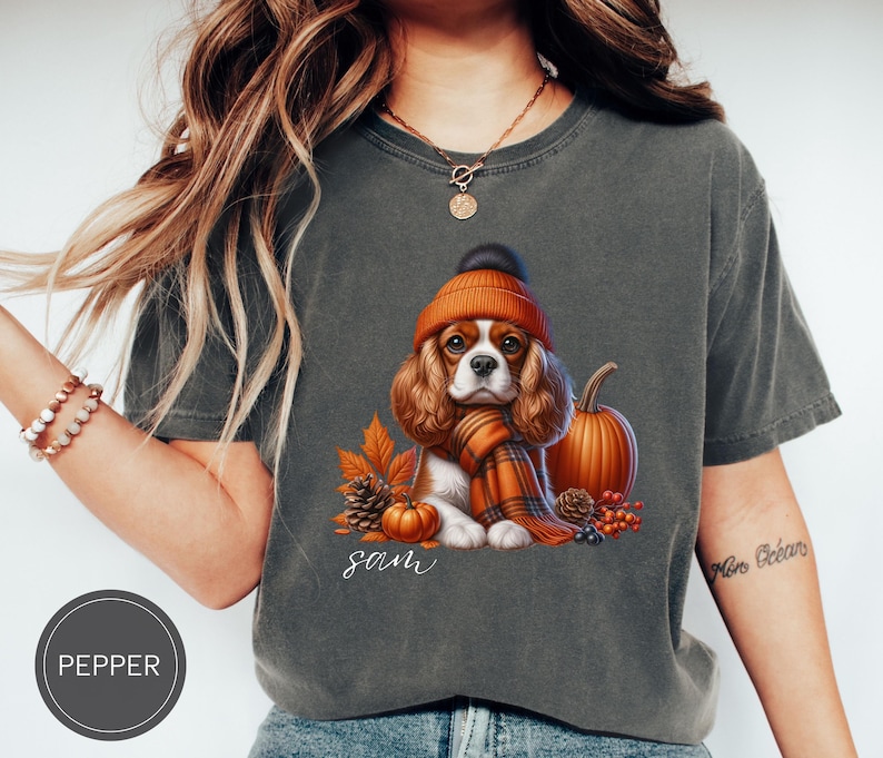 May include: A gray t-shirt with a cartoon image of a Cavalier King Charles Spaniel wearing a brown and orange hat and scarf. The dog is surrounded by fall-themed elements like pumpkins, leaves, and berries. The text "Sam" is printed below the dog.