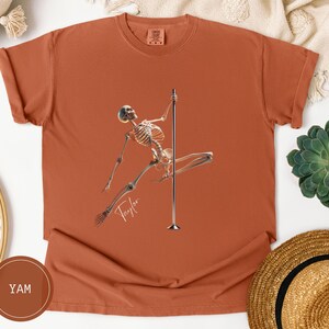 May include: Terracotta-colored t-shirt featuring a skeleton figure pole dancing. The skeleton is holding onto a silver pole with one leg extended. The word "Taylor" is written in cursive below the skeleton.