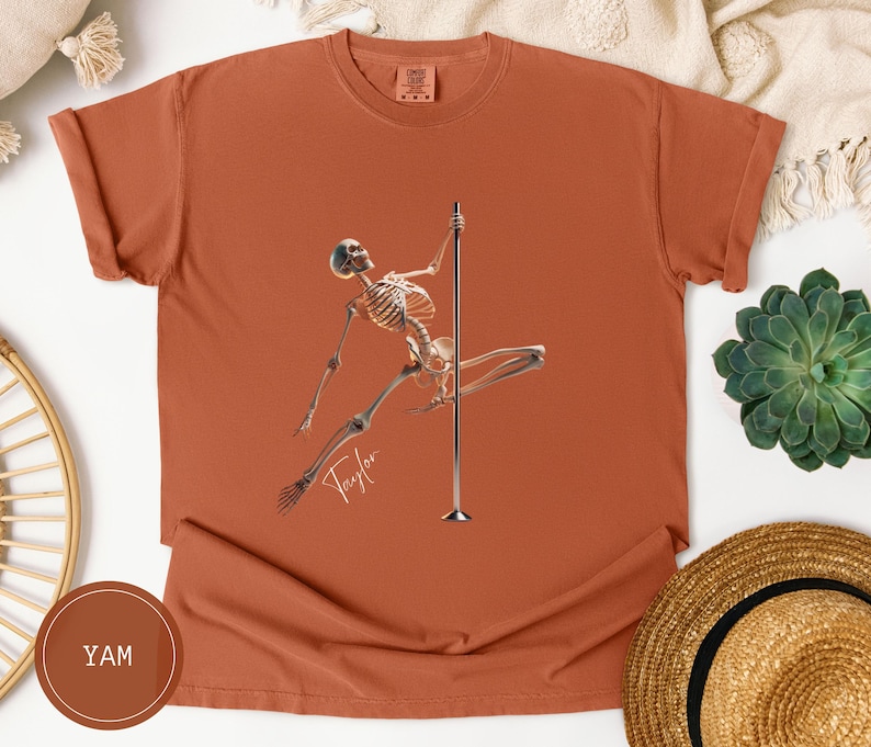 May include: Terracotta-colored t-shirt featuring a skeleton figure pole dancing. The skeleton is holding onto a silver pole with one leg extended. The word "Taylor" is written in cursive below the skeleton.