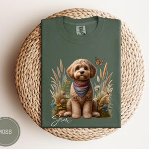 Könnte beinhalten: Ein grünes T-Shirt mit einer Illustration eines braunen und weißen Hundes. Der Hund trägt ein blaues Bandana und ist von Blumen und einem Schmetterling umgeben. Der Text "Sam" ist unter dem Hund gedruckt.