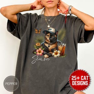 Può includere: T-shirt grigio scuro con un gatto che indossa un cappello, circondato da fiori e una bevanda. Il nome del gatto, "Simba", è scritto in corsivo. È presente anche una farfalla. La maglietta ha un aspetto vintage.