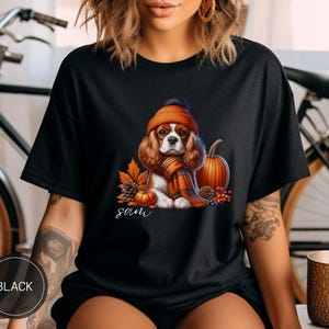 May include: Black t-shirt with a graphic of a Cavalier King Charles Spaniel wearing a pumpkin-colored hat and scarf, surrounded by fall foliage and pumpkins. The text "Sam" is printed below the dog.