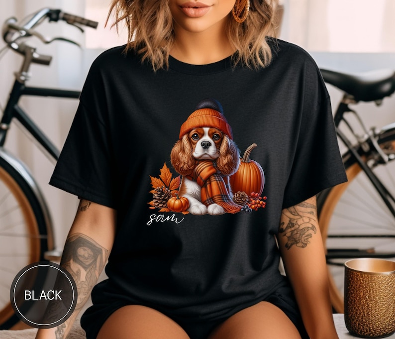 May include: Black t-shirt with a graphic of a Cavalier King Charles Spaniel wearing a pumpkin-colored hat and scarf, surrounded by fall foliage and pumpkins. The text "Sam" is printed below the dog.