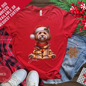 May include: Red t-shirt featuring a festive dog illustration. The dog wears a Santa hat and is wrapped in Christmas lights. Text on the shirt reads "100+ DOG BREEDS AVAILABLE OR REQUEST CUSTOM". The dog's name, "Sam", is written in cursive.