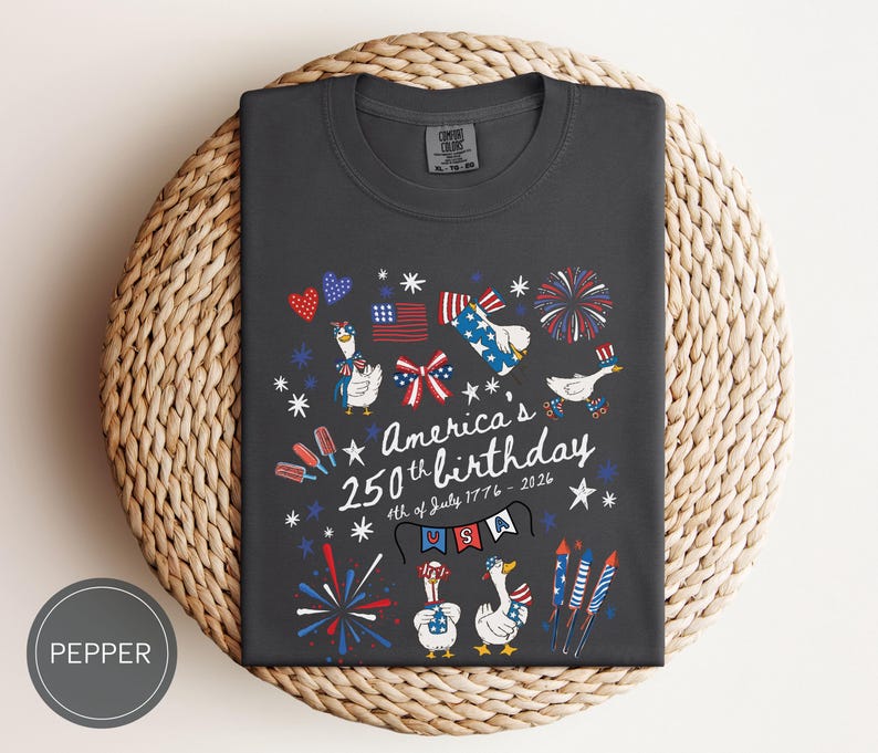 May include: Dark grey t-shirt featuring a patriotic design for America's 250th birthday. The graphic includes cartoon geese, fireworks, flags, and the text "America's 250th birthday 4th of July 1776-2026".