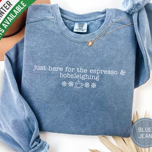 Puede incluir: Una sudadera azul vaquero con la frase "just here for the espresso & bobsleighing" bordada en blanco. El diseño incluye copos de nieve y una taza de café. Un collar de oro con un colgante está drapeado sobre la camisa.