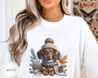 Op de afbeelding: Wit sweatshirt met een afbeelding van een teckel die een gebreide muts en sjaal draagt, omringd door dennenappels, bessen en bladeren. De tekst "Santa" is onder de hond gedrukt.
