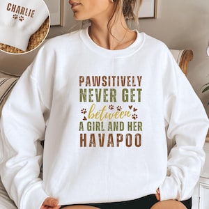 May include: White sweatshirt with the text "Pawsitively Never Get Between A Girl And Her Havapoo" in brown and green lettering. The text is surrounded by paw prints and hearts.
