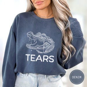 May include: A denim blue crewneck sweatshirt featuring a white line-art graphic of a crocodile head with a single tear falling from its eye. Below the illustration, the word TEARS is printed in bold, white, sans-serif capital letters.