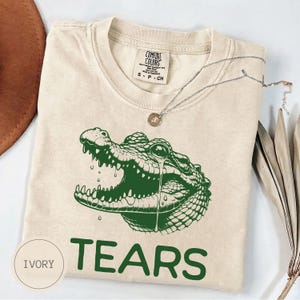 May include: An ivory short-sleeved t-shirt featuring a green graphic of a crocodile head with a single tear falling from its eye. The text "TEARS" is printed in green below the crocodile. The inner neck label reads "COMFORT COLORS S P CH".