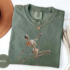 May include: Moss green t-shirt featuring a skeleton pole dancing. The graphic is in shades of brown and tan. A gold necklace with a round pendant is on the shirt. The shirt is folded and has a Comfort Colors tag.