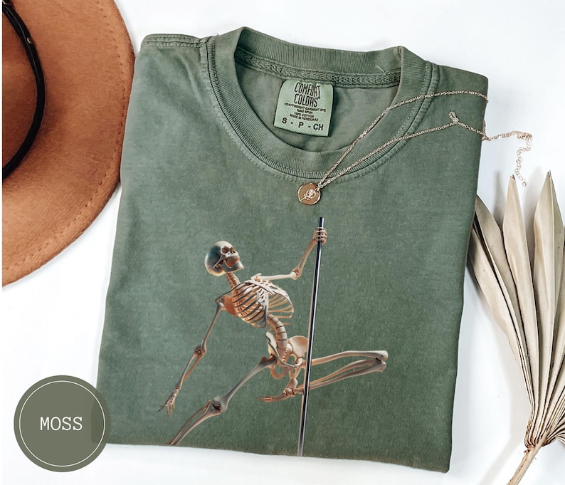 May include: Moss green t-shirt featuring a skeleton pole dancing. The graphic is in shades of brown and tan. A gold necklace with a round pendant is on the shirt. The shirt is folded and has a Comfort Colors tag.