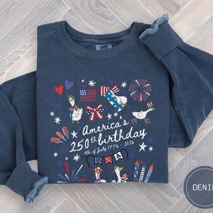 May include: A navy blue long-sleeved sweatshirt featuring a graphic commemorating America's 250th birthday. The design incorporates patriotic symbols such as flags, fireworks, and cartoon geese. The text reads "America's 250th birthday 4th of July 1776 - 2026".