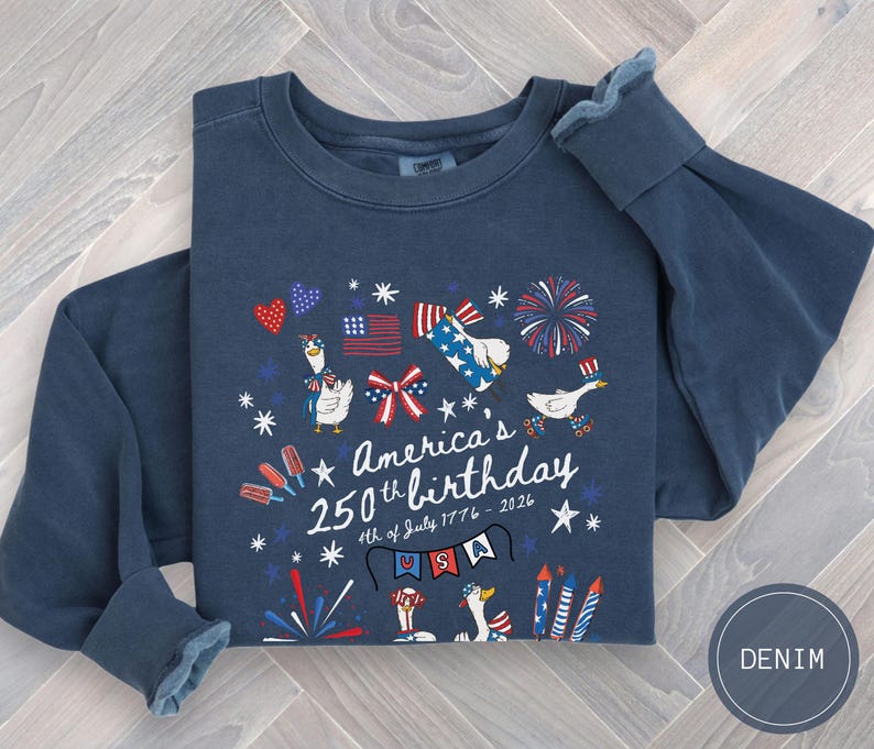 May include: A navy blue long-sleeved sweatshirt featuring a graphic commemorating America's 250th birthday. The design incorporates patriotic symbols such as flags, fireworks, and cartoon geese. The text reads "America's 250th birthday 4th of July 1776 - 2026".