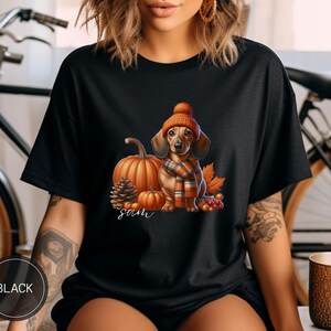 Op de afbeelding: Zwart T-shirt met een afbeelding van een teckel die een gebreide muts en sjaal draagt, omringd door pompoenen en herfstbladeren. De tekst "sam" is onder de hond gedrukt.