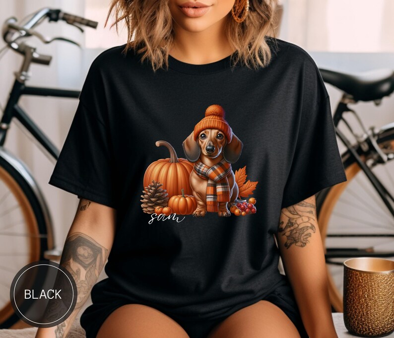 Op de afbeelding: Zwart T-shirt met een afbeelding van een teckel die een gebreide muts en sjaal draagt, omringd door pompoenen en herfstbladeren. De tekst "sam" is onder de hond gedrukt.