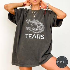 May include: A charcoal gray short-sleeve t-shirt featuring a white line-art illustration of a crocodile head with water droplets falling from its jaw. Below the graphic, the word TEARS is printed in a bold, white, sans-serif capital font.