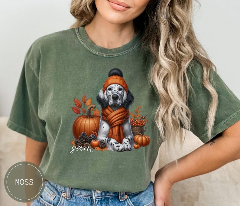 May include: A green t-shirt with a white and black dog wearing a brown and orange hat and scarf. The dog is sitting in front of a pumpkin and fall leaves. The text "Sam" is printed on the shirt.