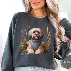 May include: A dark gray sweatshirt with a cute illustration of a fluffy white dog with a blue bandana. The dog is surrounded by flowers and grass. The text "Sam" is printed below the dog.