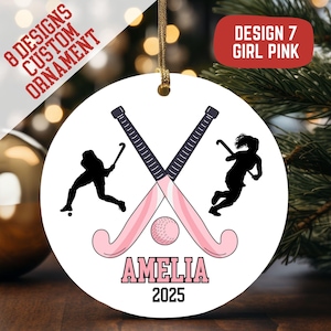 May include: A white, round ornament with a hockey theme. It features crossed pink hockey sticks, a hockey ball, and silhouettes of hockey players. The name "AMELIA" and the year "2025" are displayed. Text on the ornament reads "8 DESIGNS CUSTOM ORNAMENT" and "DESIGN 7 GIRL PINK."