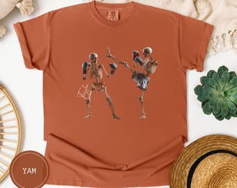 May include: A burnt orange t-shirt featuring a graphic of two skeleton boxers in action. The skeletons wear boxing gloves and shorts. The word "Taylor" is written in cursive on the shirt.