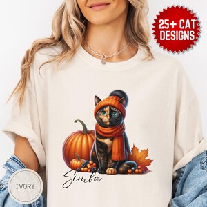 Può includere: T-shirt color avorio con un gatto che indossa un cappello e una sciarpa arancioni, con una zucca e foglie autunnali. Il gatto è nero, marrone e arancione. Il testo "Simba" è scritto in corsivo. La maglietta ha anche il testo "25+ CAT DESIGNS".