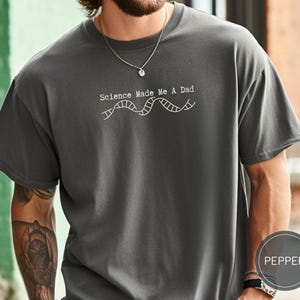 Op de afbeelding: Donkergrijze t-shirt met de witte tekst "Science Made Me A Dad" en een DNA-strengafbeelding. Het t-shirt heeft een ronde hals en korte mouwen.