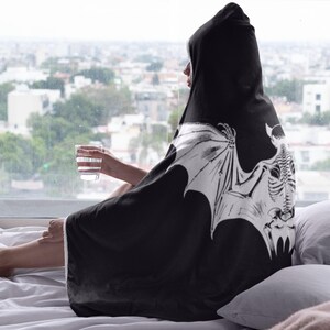 May include: A black hooded blanket with a white skeletal bat design. The blanket is draped over a person sitting on a bed.