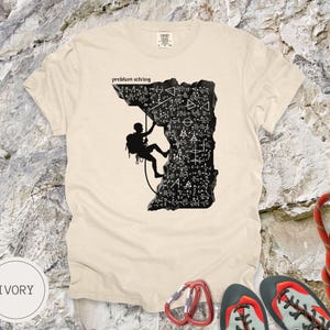 May include: Ivory-coloured t-shirt with a black graphic of a climber on a rock face covered in mathematical symbols. The text "problem solving" is above the climber. Climbing shoes, a carabiner, and a rope are visible.