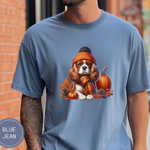May include: A blue t-shirt with a cartoon image of a Cavalier King Charles Spaniel wearing a brown and orange knit hat and scarf. The dog is sitting next to a small orange pumpkin and autumn leaves.