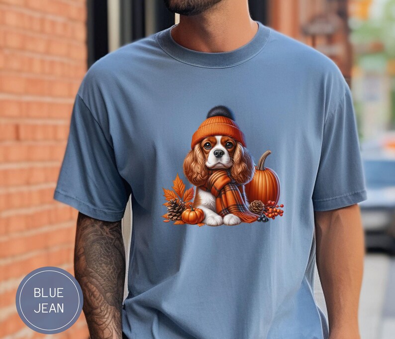 May include: A blue t-shirt with a cartoon image of a Cavalier King Charles Spaniel wearing a brown and orange knit hat and scarf. The dog is sitting next to a small orange pumpkin and autumn leaves.