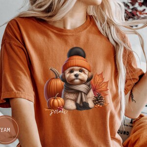 Puede incluir: Una camiseta marrón con un lindo perro que lleva un sombrero y una bufanda marrones, rodeado de calabazas y hojas de otoño. El texto "Sarah" está impreso en la camiseta.