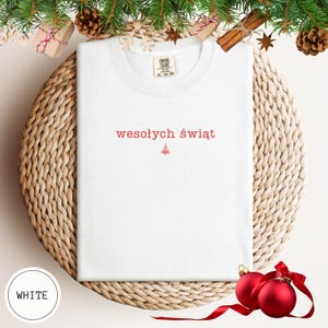 Może przedstawiać: Biały t-shirt z kr&oacute;tkim rękawem z czerwonym napisem "wesołych świąt" wyśrodkowanym na klatce piersiowej, kt&oacute;remu towarzyszy mała czerwona grafika choinki umieszczona bezpośrednio pod tekstem. Koszulka posiada okrągły dekolt oraz metkę.