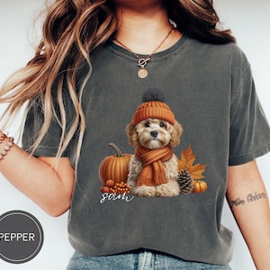 Puede incluir: Una camiseta gris con un lindo perro que lleva un gorro y una bufanda marrones y naranjas. El perro está rodeado de calabazas y hojas de otoño. El texto "Sam" está impreso en la camiseta.