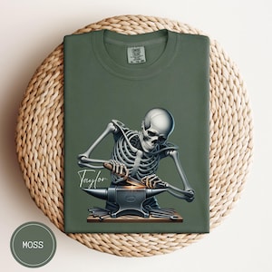 May include: Moss green t-shirt featuring a detailed illustration of a skeleton blacksmith working on an anvil. The skeleton is depicted with a hammer and a glowing hot metal. The name "Taylor" is written in cursive. The shirt is made of a soft material.