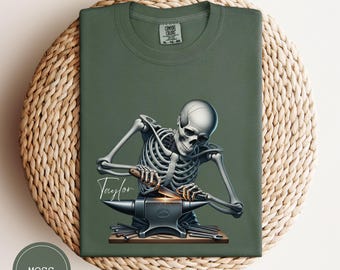 May include: Moss green t-shirt featuring a detailed illustration of a skeleton blacksmith working on an anvil. The skeleton is depicted with a hammer and a glowing hot metal. The name "Taylor" is written in cursive. The shirt is made of a soft material.