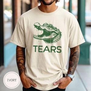 May include: An ivory short-sleeved t-shirt featuring a dark green graphic of a crocodile with an open mouth and a single tear falling from its eye. Below the crocodile illustration, the word TEARS is printed in a bold, dark green sans-serif font.